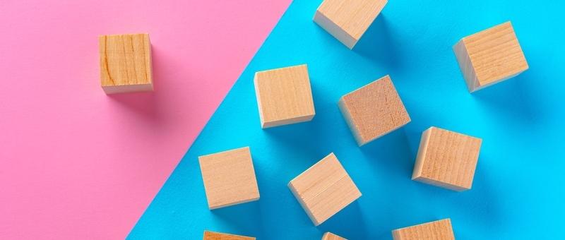 Wooden blocks against a split pink and blue background. One block against pink, with multiple blocks against blue.