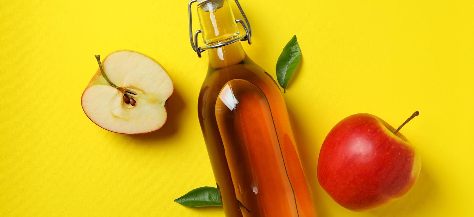 Glass bottle of apple cider vinegar with fresh red apple, apple slice, and green leaves on bright yellow background.