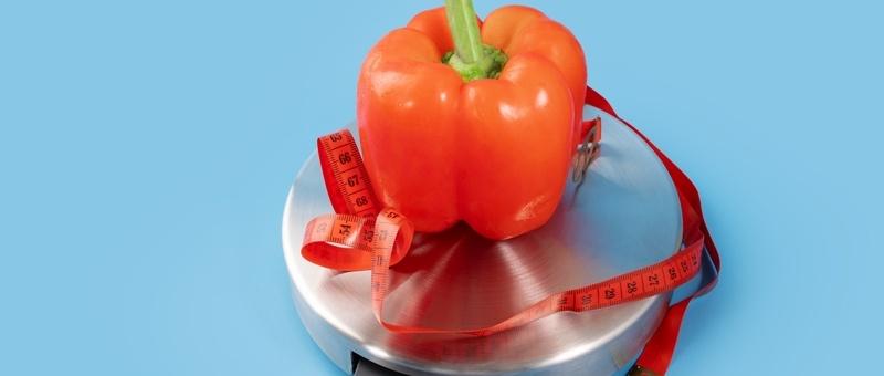 Red bell pepper on silver weighing scale surrounded by red measuring tape against blue background.