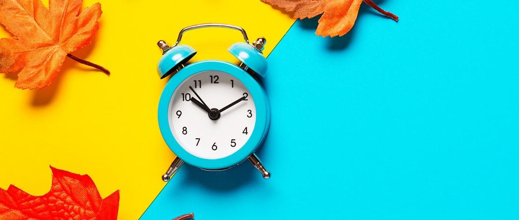 Clock on two tone colour yellow and light blue background decorated with colourful fallen leaves.